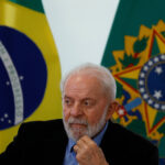 Homem de cabelos e barba brancos veste terno preto e camisa azul, sentado com mão no queixo. Ao fundo, bandeira do Brasil e símbolo nacional em fundo verde.