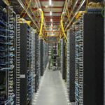 A imagem mostra uma sala de servidores com fileiras de equipamentos de computação. Os servidores estão organizados em racks altos, com cabos azuis e amarelos conectando-os. No fundo, há uma escada e uma pessoa em pé, possivelmente trabalhando nos equipamentos. A iluminação é artificial e o ambiente é bem organizado.