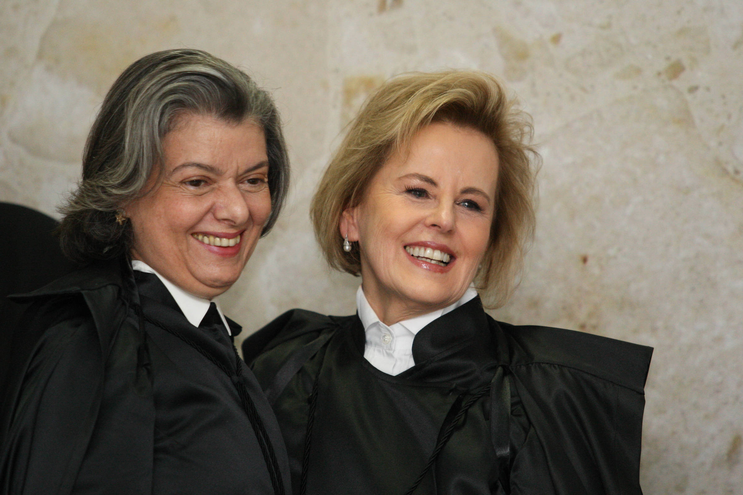 Duas mulheres vestindo togas pretas e camisas brancas posam sorrindo lado a lado contra parede clara. Ambas têm cabelos curtos, uma com cabelo grisalho e outra loiro claro.