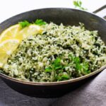A imagem mostra um prato de arroz verde preparado com folhas picadas misturadas aos grãos, decorado com fatias de limão e salsinha fresca. Pela aparência, trata-se de um arroz com ora-pro-nóbis