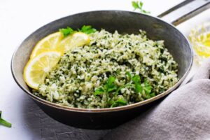 A imagem mostra um prato de arroz verde preparado com folhas picadas misturadas aos grãos, decorado com fatias de limão e salsinha fresca. Pela aparência, trata-se de um arroz com ora-pro-nóbis