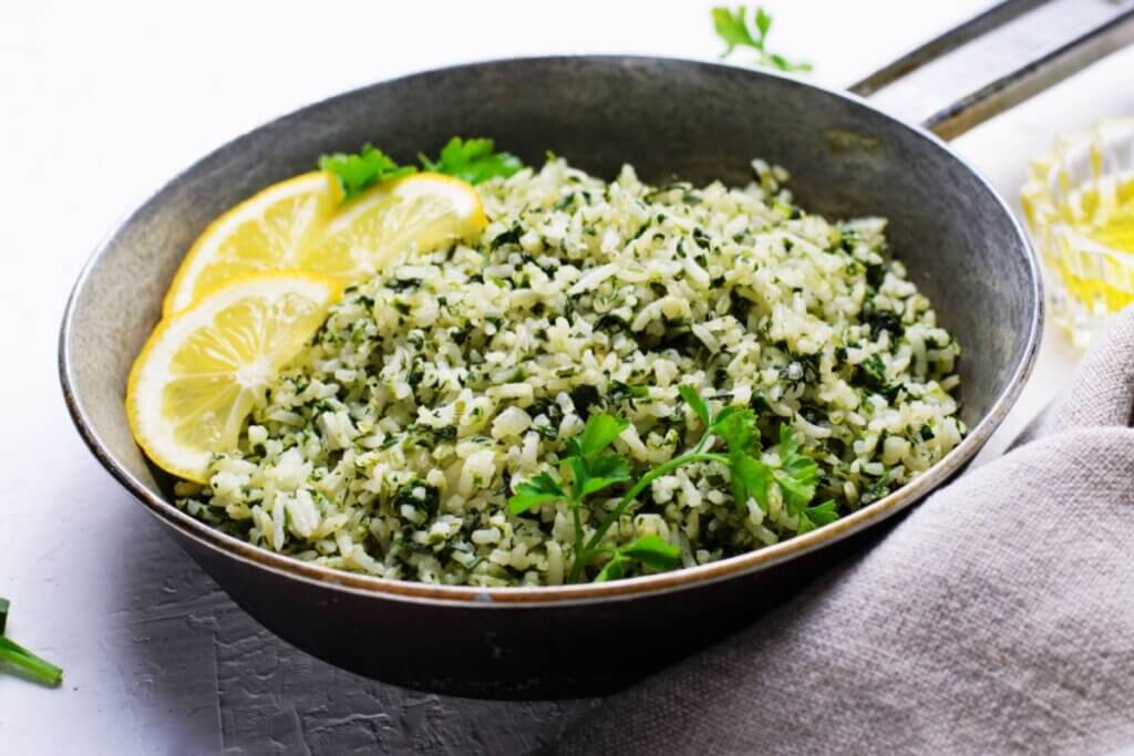 A imagem mostra um prato de arroz verde preparado com folhas picadas misturadas aos grãos, decorado com fatias de limão e salsinha fresca. Pela aparência, trata-se de um arroz com ora-pro-nóbis