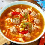 Tigela de sopa com almôndegas de peru, grão-de-bico, tiras de massa, cenoura e pimentão vermelho em caldo avermelhado, finalizada com dill