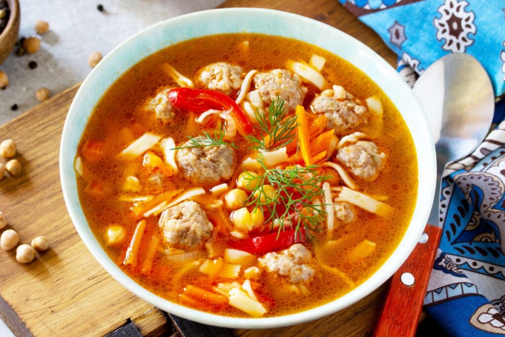 Tigela de sopa com almôndegas de peru, grão-de-bico, tiras de massa, cenoura e pimentão vermelho em caldo avermelhado, finalizada com dill