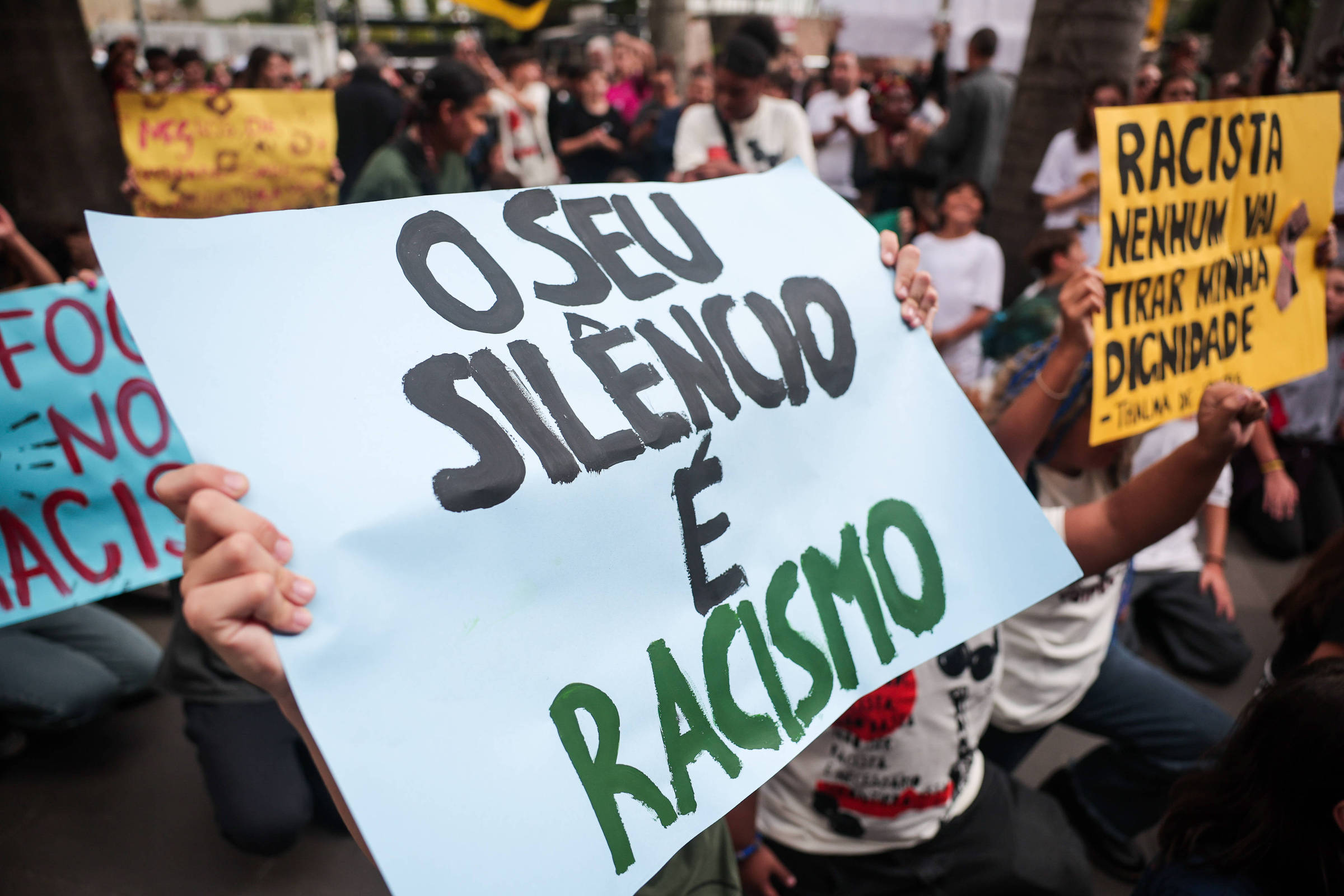 A imagem mostra um grupo de pessoas participando de um protesto. Em primeiro plano, uma pessoa segura um cartaz azul com a frase