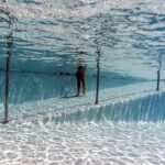 Duas pessoas caminham sobre passarela submersa em piscina com água clara. Estrutura metálica suporta passarela, e reflexos de luz criam padrões no fundo da piscina.