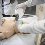 Profissionais de saúde praticam reanimação cardiopulmonar em manequim adulto em ambiente hospitalar. Um aplica compressões torácicas enquanto outro utiliza bolsa de ventilação manual para insuflar ar no manequim.