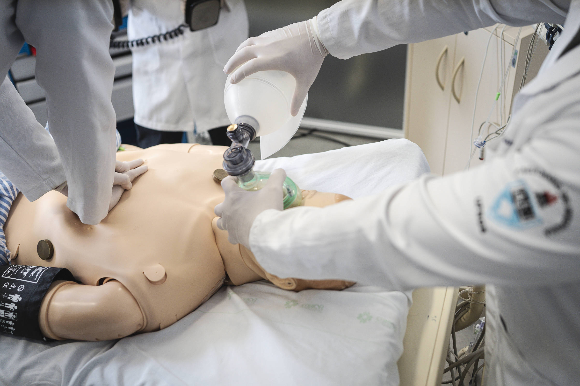 Profissionais de saúde praticam reanimação cardiopulmonar em manequim adulto em ambiente hospitalar. Um aplica compressões torácicas enquanto outro utiliza bolsa de ventilação manual para insuflar ar no manequim.