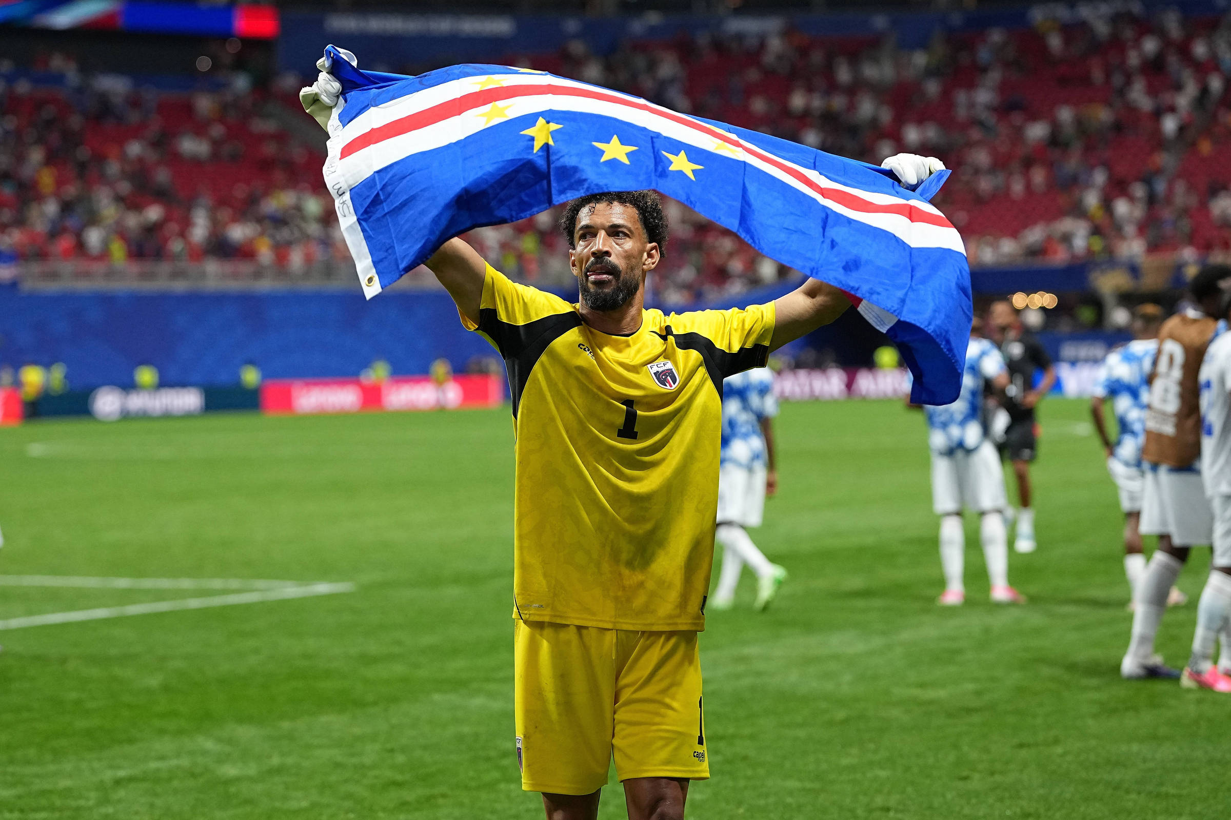 Jogador de futebol em uniforme amarelo levanta a bandeira de Cabo Verde acima da cabeça no campo de jogo. Ao fundo, outros jogadores e arquibancadas com público.