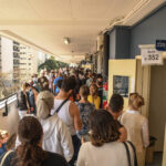 Um corredor lotado de filas de pessoas, uma delas posicionada diante de uma porta bege aberta. Prédios aparecem pela sacada do prédio