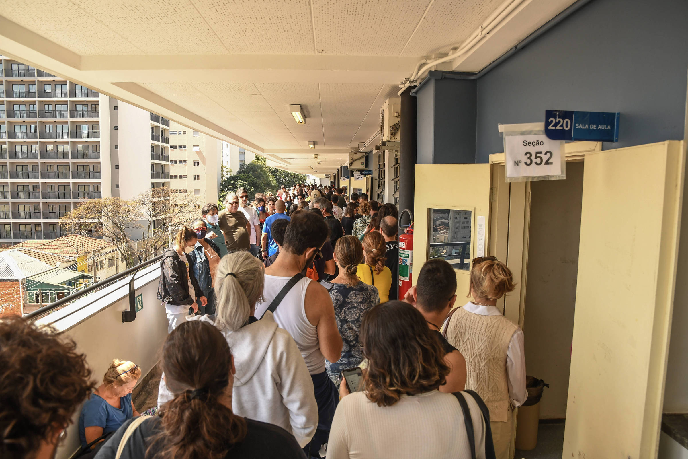 Um corredor lotado de filas de pessoas, uma delas posicionada diante de uma porta bege aberta. Prédios aparecem pela sacada do prédio