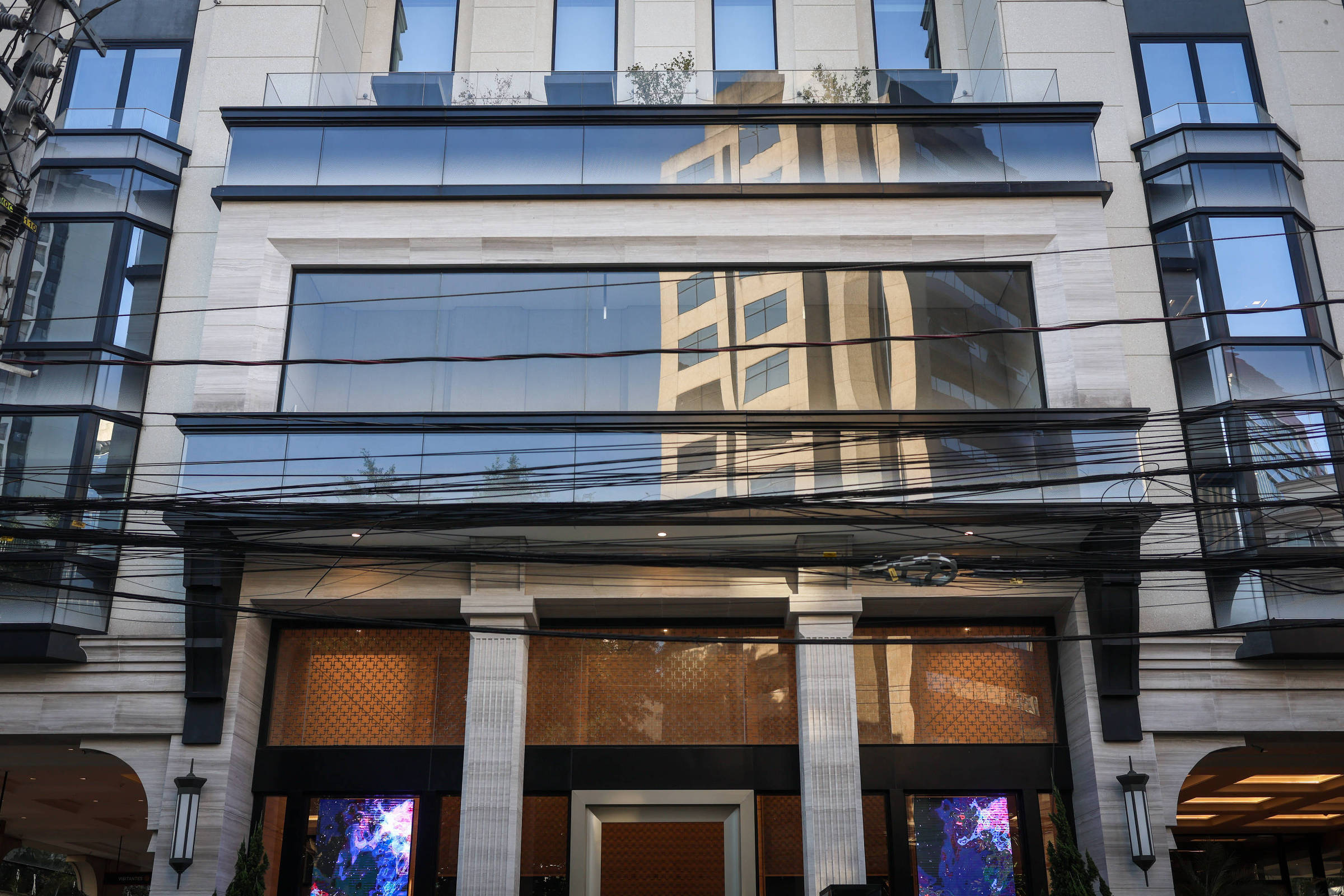 Fachada de edifício comercial com grandes janelas espelhadas que refletem outro prédio alto. A entrada tem colunas claras e painéis decorativos, com duas telas digitais coloridas nas laterais. Fios elétricos cruzam horizontalmente em frente ao prédio.