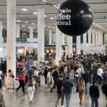 A imagem mostra uma grande multidão em um evento, possivelmente um festival de café, com várias barracas e estandes ao fundo. Um balão preto com a inscrição