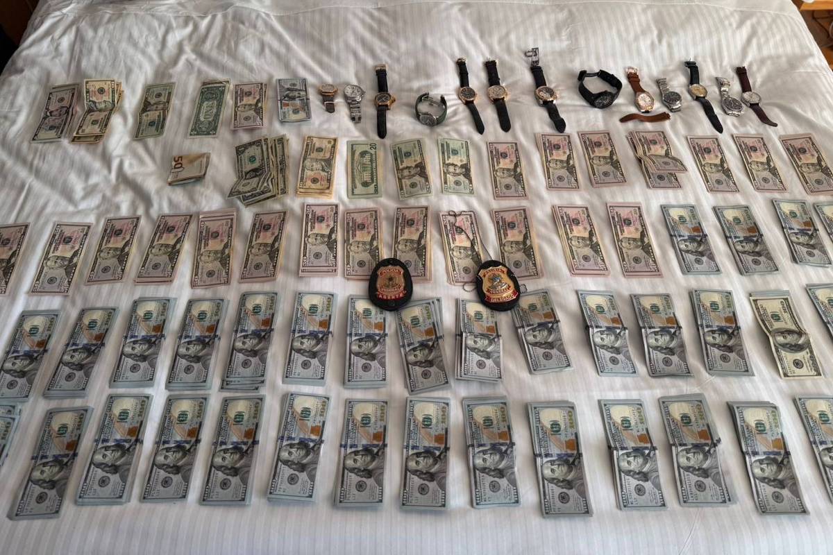 Diversas cédulas de dólares organizadas em fileiras sobre uma cama branca, acompanhadas por vários relógios de pulso e dois distintivos policiais no centro da disposição.