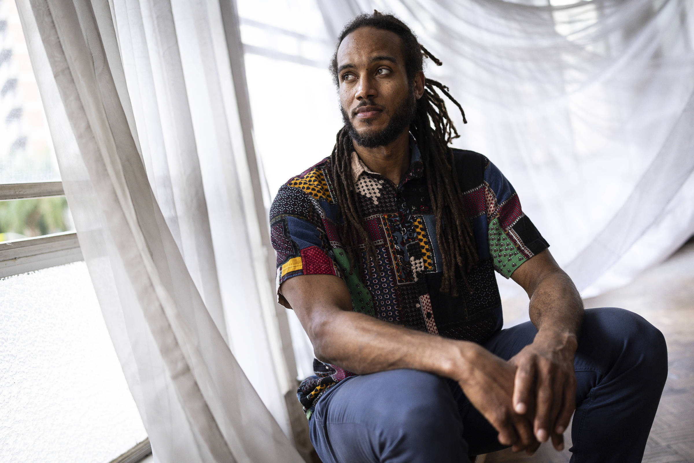 Homem com cabelos longos presos em dreadlocks está sentado próximo a uma janela com cortinas brancas translúcidas. Ele veste camisa colorida de padrão geométrico e calça azul, olhando para o lado direito da imagem.
