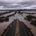 A imagem mostra uma área urbana severamente inundada, com grandes extensões de água cobrindo a vegetação e as estradas. No centro, há uma estrada que se estende em direção a um ponto onde a água parece mais profunda. Algumas árvores e arbustos são visíveis, mas a maior parte da paisagem está submersa. O céu está nublado, indicando condições climáticas adversas.