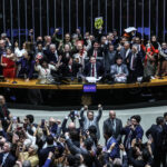 Grupo numeroso de deputados em pé atrás da mesa principal do plenário, muitos com os punhos erguidos em comemoração. Na frente, público e outros parlamentares também registram o momento com celulares. Ambiente interno com iluminação artificial e estrutura típica de assembleia legislativa.
