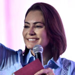 Uma mulher está em um palco, segurando um microfone com a mão direita e um caderno ou livro com a mão esquerda. Ela está sorrindo e acenando com a mão, vestindo uma camisa azul com detalhes. O fundo é de cor rosa, sugerindo um ambiente de evento ou apresentação.