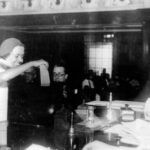 A imagem mostra uma cena de tribunal, onde uma mulher com um vestido claro e um chapéu está em pé, levantando um documento. À sua frente, um homem de óculos está sentado, enquanto outro homem mais velho, com cabelo grisalho, observa a cena. O ambiente é formal, com mesas e documentos visíveis, e há várias pessoas sentadas ao fundo, assistindo ao que acontece.