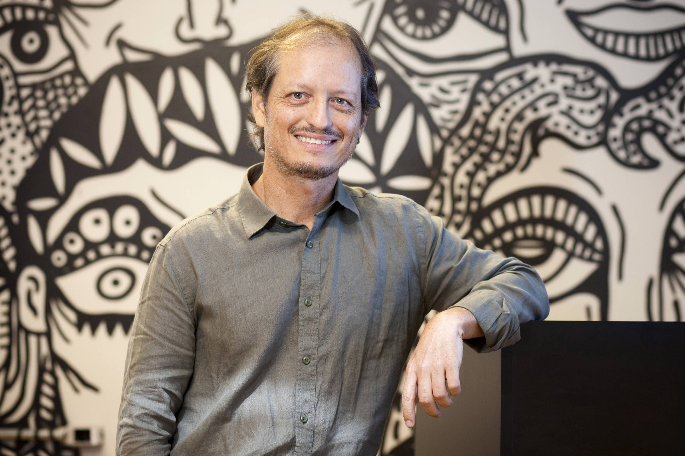 Um homem sorridente está posando para a foto, com uma camisa de botão cinza. Ele está apoiado em uma superfície preta e tem os braços cruzados. Ao fundo, há uma parede decorada com um mural artístico em preto e branco, apresentando rostos e elementos gráficos. A iluminação é suave, destacando o homem e o mural.
