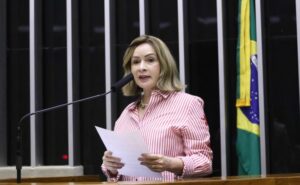Deputada Socorro Neri fala ao microfone. Ela é uma mulher branca, tem o cabelo liso, loiro, na altura dos ombros, veste uma camisa listrada de vermelho e branco e segura um papel