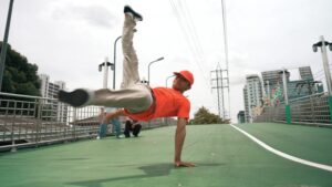 Um dançarino de boné e camiseta vermelhos executa um movimento de breakdance, apoiado em uma das mãos e com as pernas erguidas. A apresentação ocorre em uma passarela urbana de piso verde, cercada por grades, postes, prédios e fios de alta tensão, sob o céu nublado