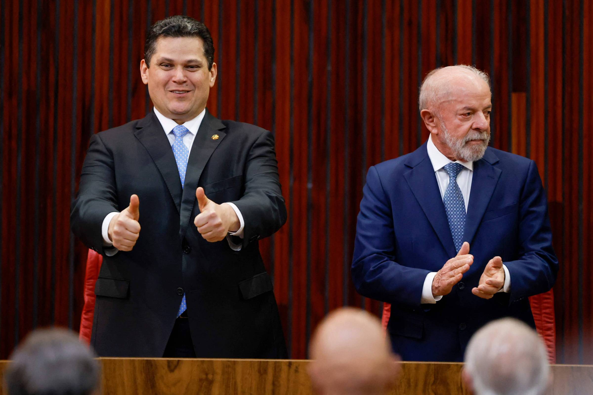 Dois homens de terno estão em pé em um ambiente formal com fundo de painéis de madeira escura. O homem à esquerda sorri e faz sinal de positivo com as duas mãos, enquanto o homem à direita, de cabelos brancos e barba, aplaude olhando para o lado. Três pessoas estão parcialmente visíveis em primeiro plano, de costas.