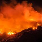 Fogo intenso consome vegetação em área de serra durante a noite, com grandes nuvens de fumaça alaranjada e chamas visíveis ao longo do terreno irregular.