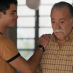 Homem jovem com camiseta marrom clara toca o ombro de homem idoso de camisa xadrez bege, ambos em ambiente interno com janela ao fundo.