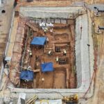 Vista aérea mostra canteiro de obras com escavações retangulares cercadas por tapumes. Cinco trabalhadores com coletes refletores estão no local, com duas tendas azuis para sombra. Equipamentos de construção, incluindo guindaste, estão posicionados ao redor da área.