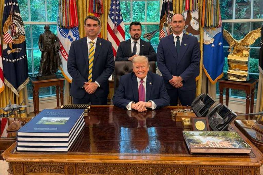 Homem sentado na mesa presidencial do Salão Oval, com três homens em pé atrás dele. Ao fundo, janelas com cortinas douradas, várias bandeiras americanas e estátuas decorativas.