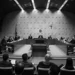 Plenário do Supremo Tribunal Federal com ministros sentados em semicírculo ao redor de mesa central. Público observa a sessão em cadeiras na parte inferior da imagem. Paredes com textura e símbolos oficiais ao fundo.