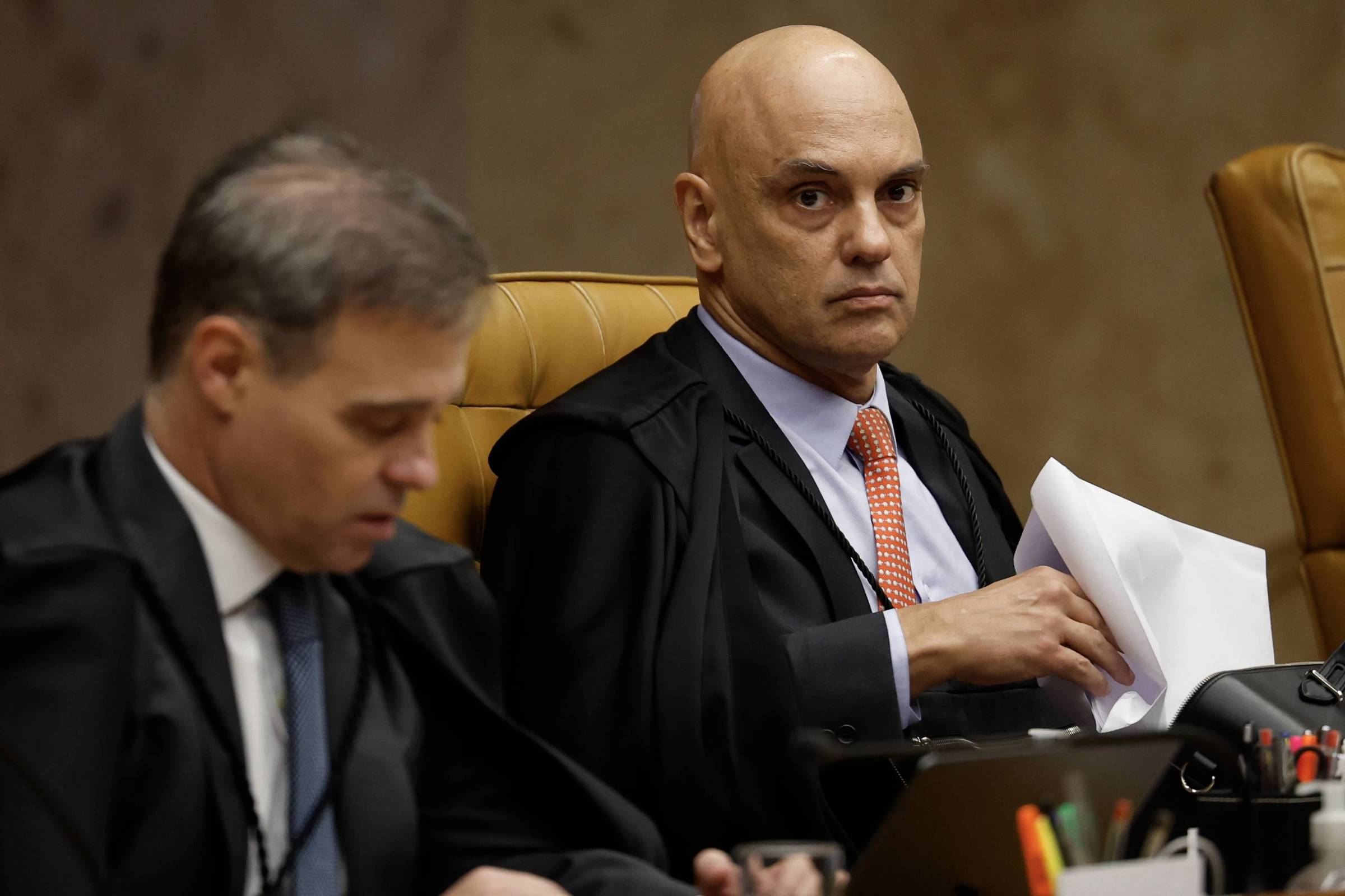 Dois homens sentados lado a lado em cadeiras de couro marrom, ambos vestindo togas pretas. O homem à direita, calvo, olha diretamente para a câmera segurando papéis, enquanto o homem à esquerda, com cabelo grisalho, está focado em documentos à sua frente. O ambiente sugere uma sessão formal, possivelmente judicial.