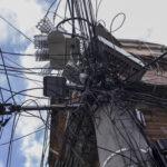 Poste de concreto com grande quantidade de fios elétricos e de telecomunicações entrelaçados e desorganizados, conectados a equipamentos e caixas instaladas no poste. Edifícios residenciais aparecem ao fundo sob céu parcialmente nublado.