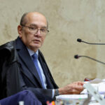 Senhor com óculos e terno azul escuro, usando toga preta, sentado em cadeira amarela em ambiente de tribunal. Ele segura papéis e está diante de microfones e objetos de escritório sobre a mesa.