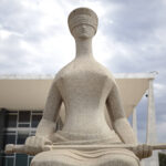 Estátua de pedra da Justiça cega sentada, segurando uma espada horizontalmente, com prédio moderno ao fundo e céu nublado.