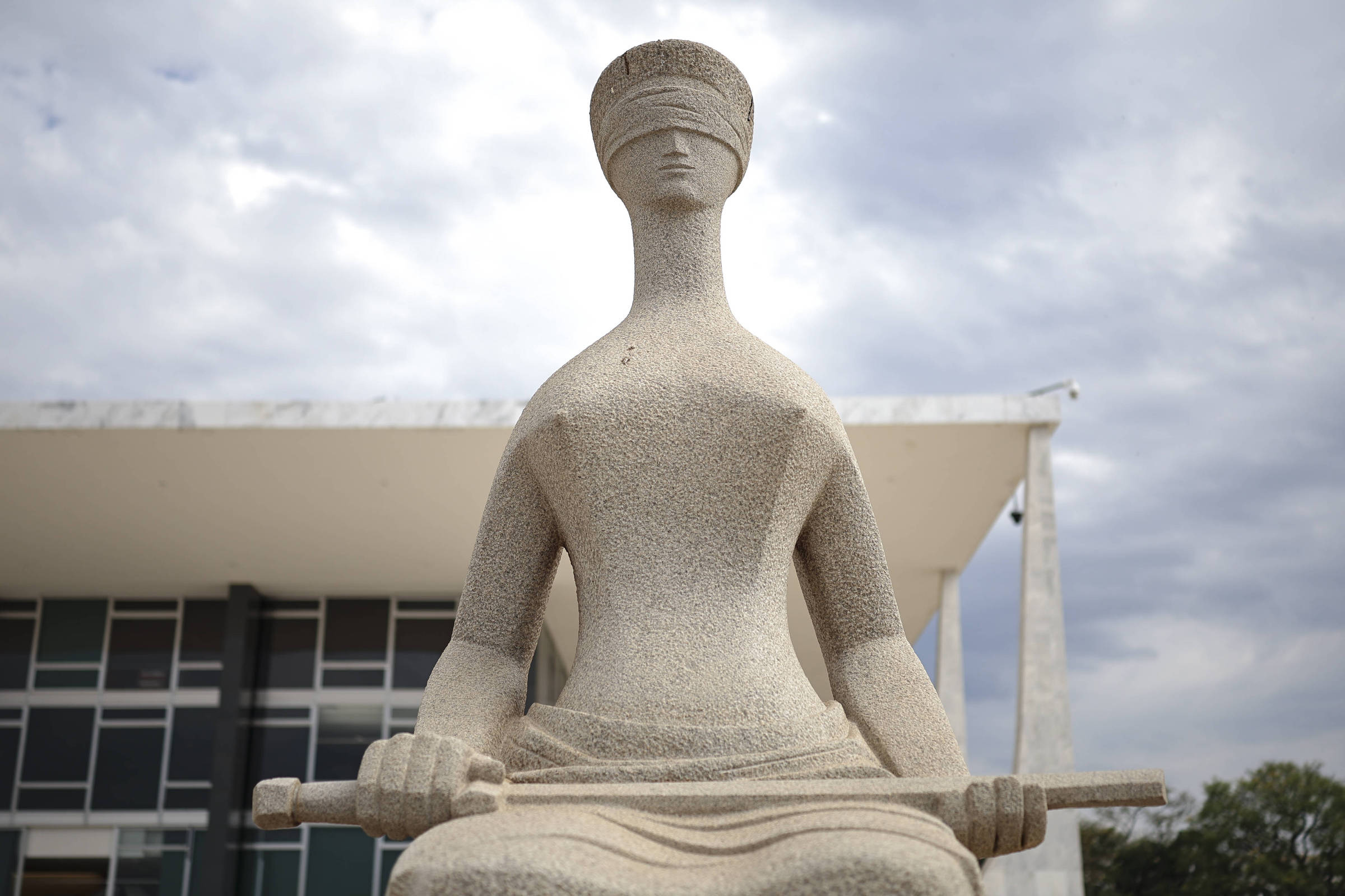 Estátua de pedra da Justiça cega sentada, segurando uma espada horizontalmente, com prédio moderno ao fundo e céu nublado.