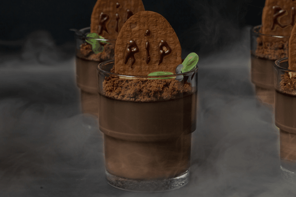 Sobremesa de Halloween em um copo de vidro, decorada como um pequeno túmulo, em destaque no centro, com outros copos iguais desfocados ao fundo. O copo tem uma camada de creme de chocolate claro, coberta por cacau em pó imitando terra, com um biscoito de chocolate em formato de lápide escrito