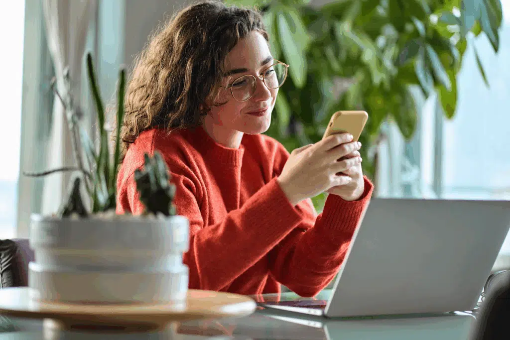 Mulher de óculos e suéter alaranjado sorri enquanto digita no celular, sentada em uma mesa de trabalho com um notebook aberto e plantas ao fundo.