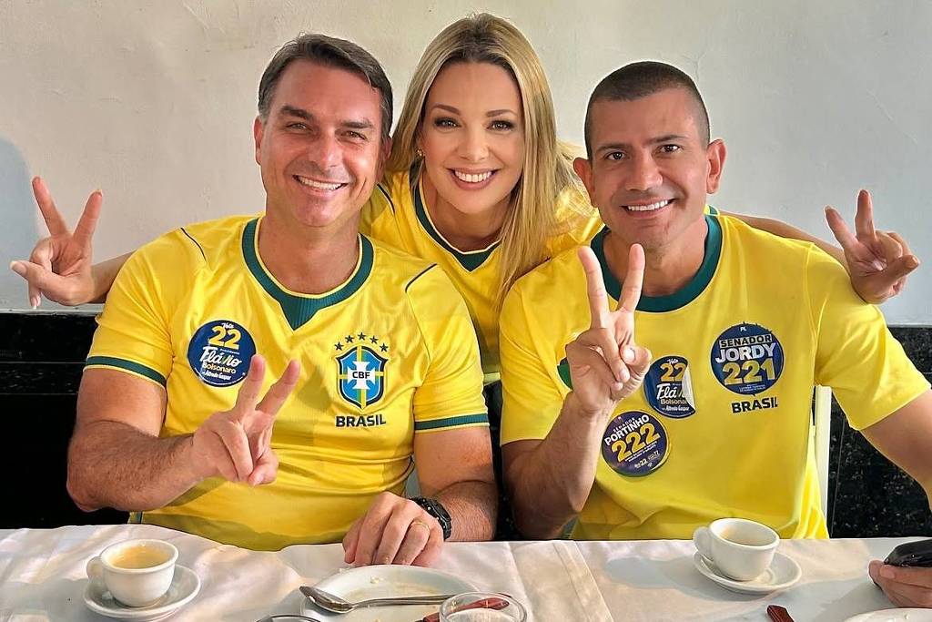 Três adultos sentados lado a lado em uma mesa de restaurante, todos vestindo camisas amarelas da seleção brasileira. Dois homens estão nas extremidades e uma mulher no centro, todos sorrindo e fazendo sinal de paz com as mãos. Na mesa há xícaras de café, copo de água, pratos e talheres.