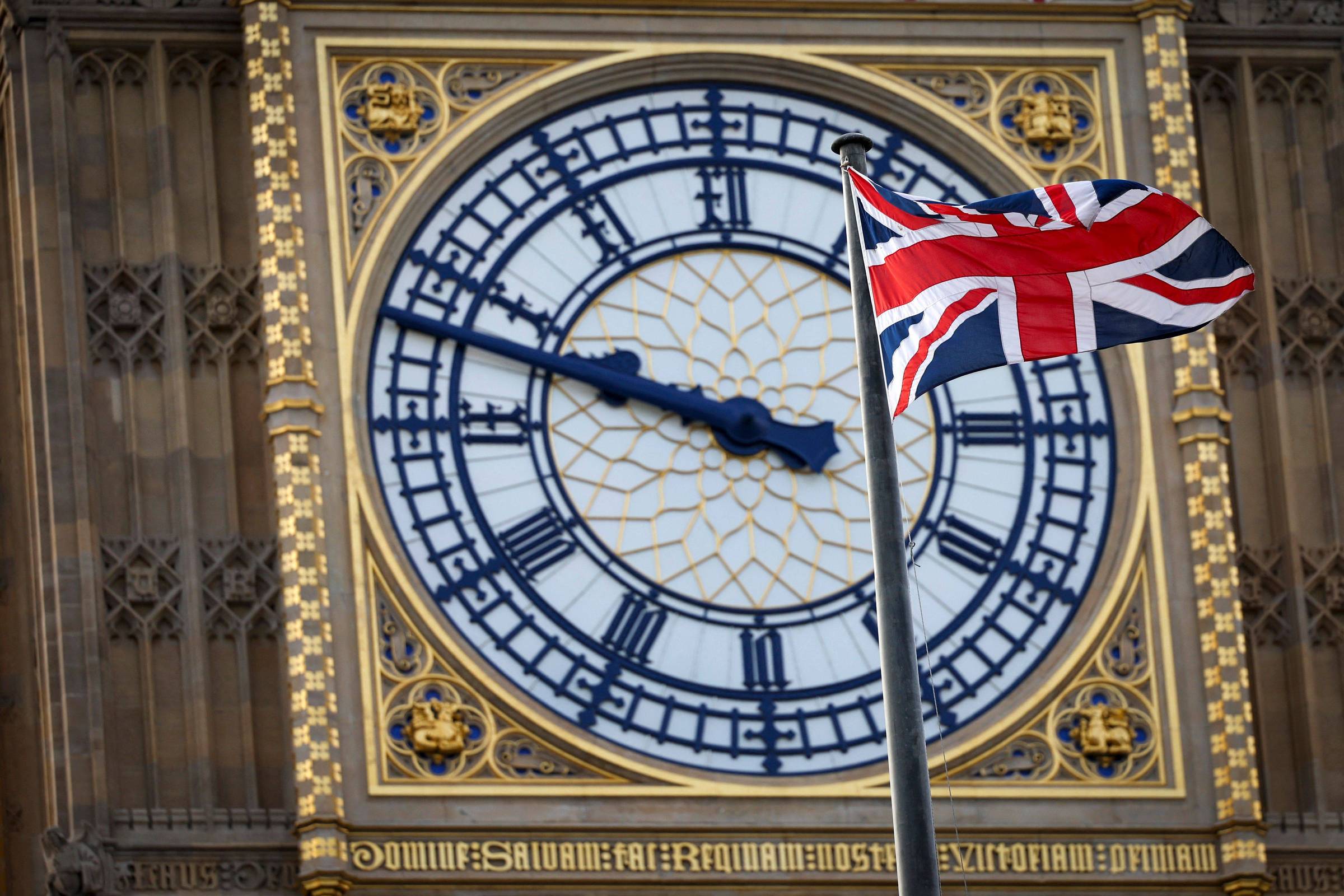 Relógio do Big Ben mostra 9h48 com detalhes dourados e números romanos. Bandeira do Reino Unido tremula em mastro à frente do relógio.