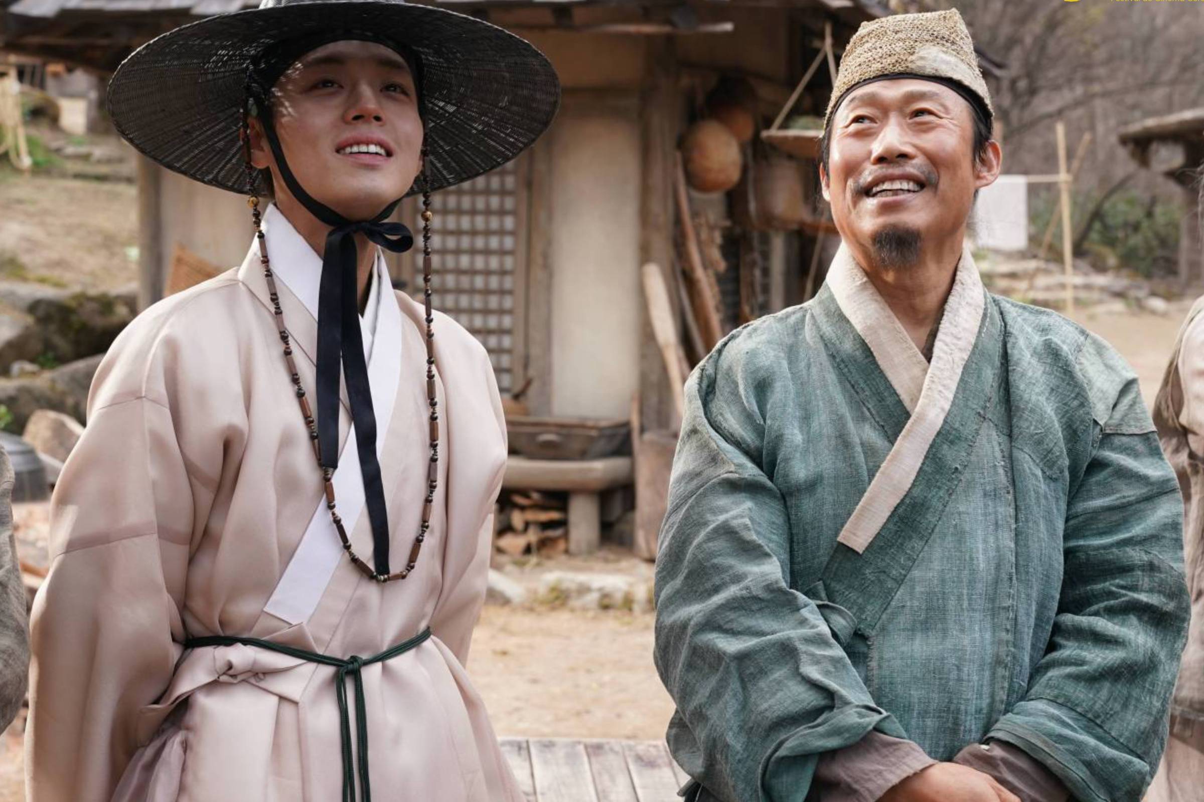 Dois homens vestindo roupas tradicionais coreanas sorriem em área externa com casa de madeira ao fundo. Um usa chapéu preto largo e o outro, gorro marrom.