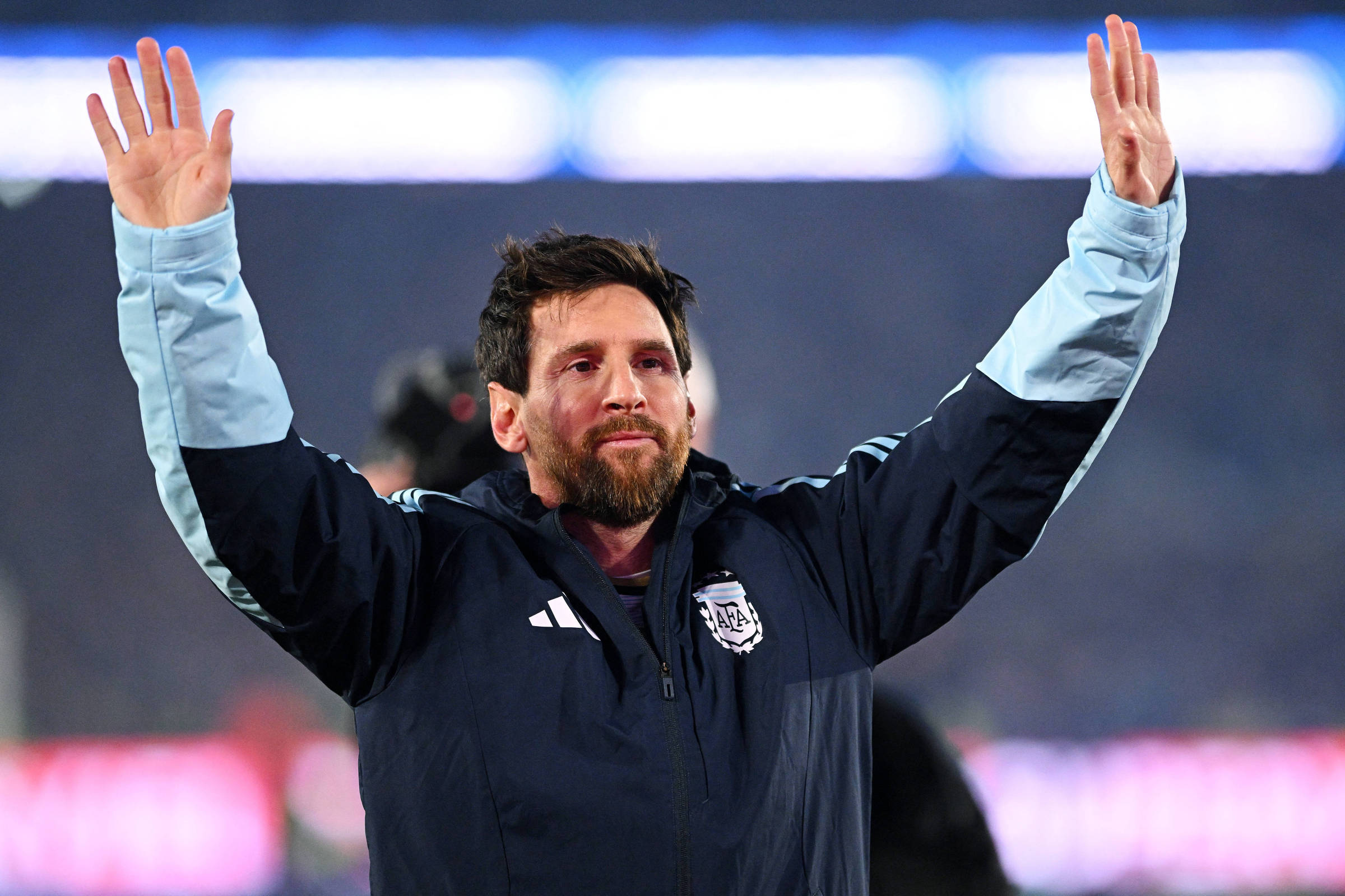 Jogador de futebol com jaqueta azul da seleção argentina levanta ambas as mãos em gesto de saudação ou agradecimento, com estádio desfocado ao fundo.