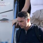 Homem com mochila preta desce escada de avião segurando corrimão. Mulher atrás veste regata branca e carrega casaco bege. Avisos em inglês e espanhol indicam para não tocar ou segurar corrimão.