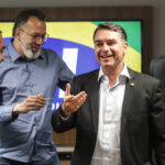 Quatro homens em trajes formais e casuais conversam e sorriem em ambiente interno. Ao fundo, tela exibe cores verde, amarela e azul, sugerindo bandeira do Brasil.