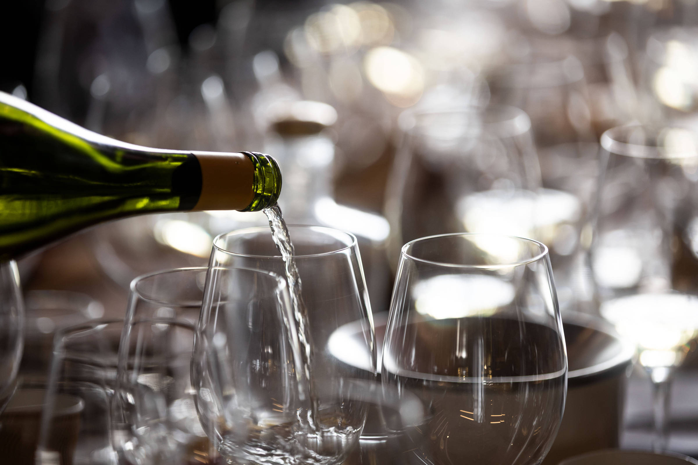 Vinho branco é servido de garrafa em taça de vidro transparente. Ao redor, várias taças e pratos estão dispostos em mesa com iluminação suave e fundo desfocado.